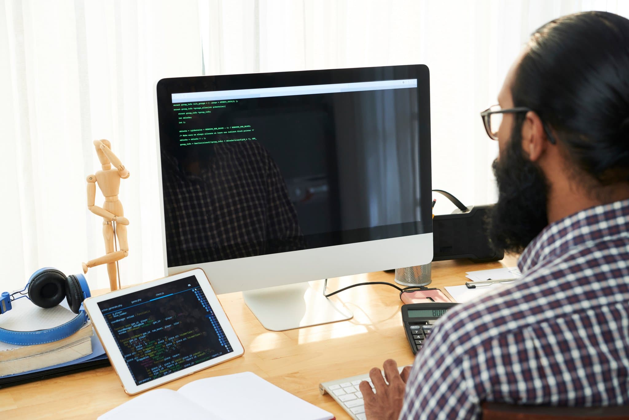 A software developer or IT engineer working at a desk with multiple devices, including a desktop computer and tablet both displaying code with syntax highlighting. The person is wearing glasses and a plaid shirt, seated at a wooden desk in what appears to be a modern office environment.